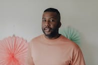 A bearded man in a pink shirt poses against a pastel decorated background.